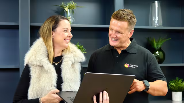 Two colleagues standing and smiling while discussing something on a laptop.
